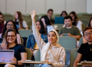 Diverse group of students in a lecture hall. Student in Hijab raising her hand to ask a question.